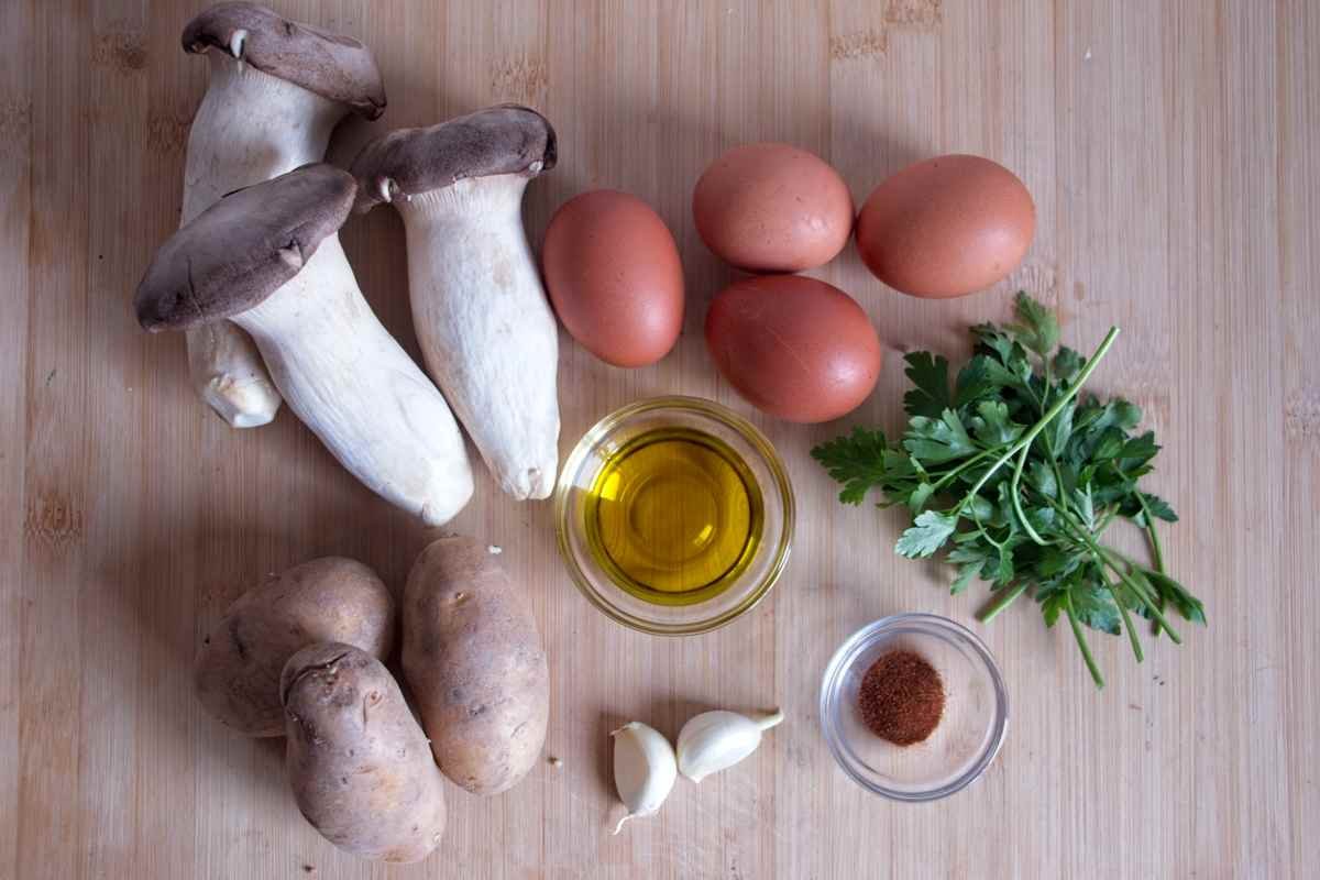 Ingredients for king oyster mushroom breakfast hash laid out on a wooden board, including mushrooms, eggs, potatoes, garlic, olive oil, herbs, and smoked paprika.