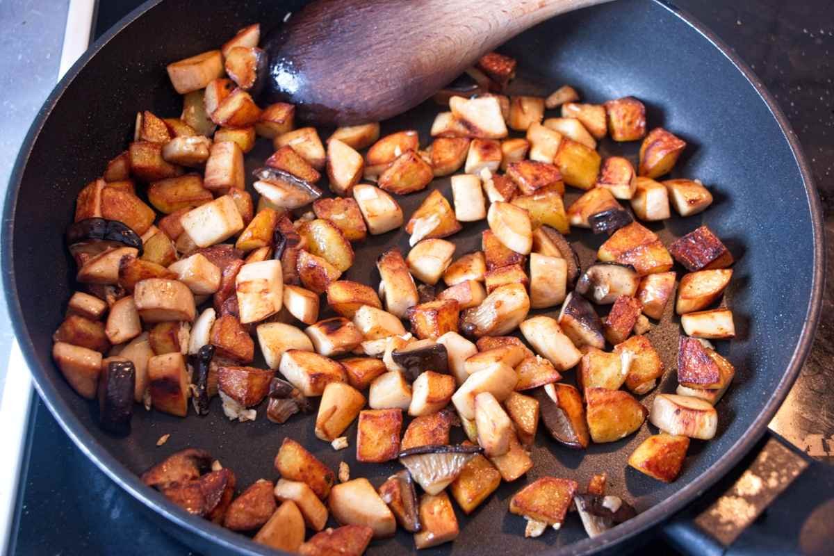 Diced potatoes and sliced mushrooms browning together in a skillet with a wooden spoon.
