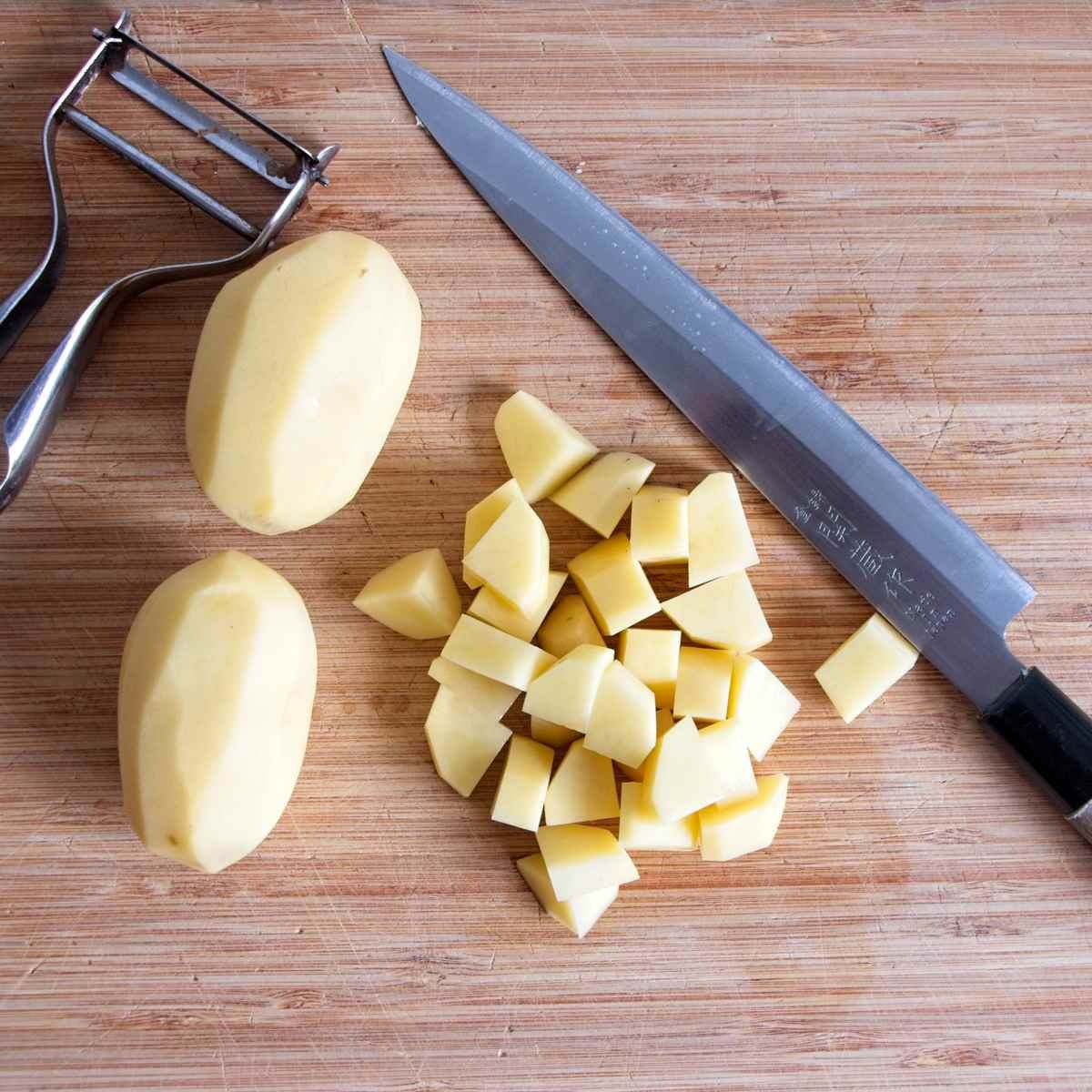 Peeled potatoes and diced cubes on a wooden board next to a knife and peeler.