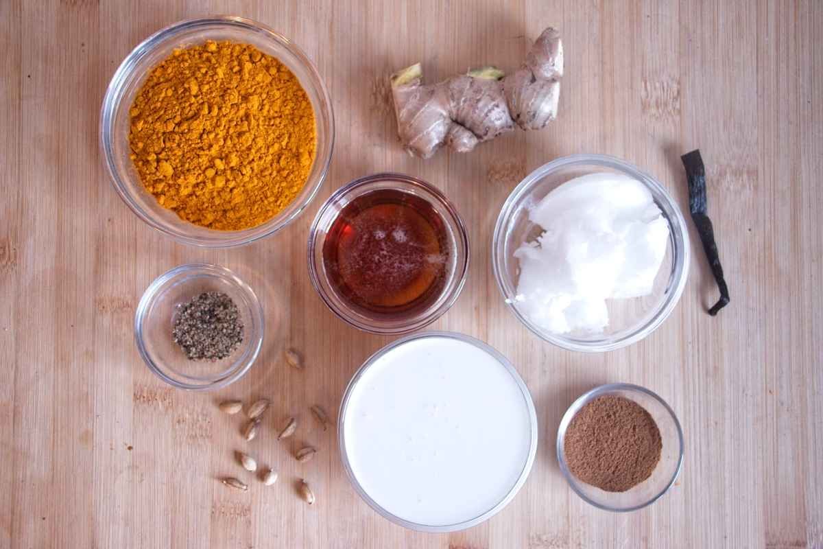 Ingredients for golden milk concentrate arranged on a wooden board, including turmeric, fresh ginger, coconut milk, coconut oil, maple syrup, cinnamon, cardamom, black pepper, and vanilla pod.
