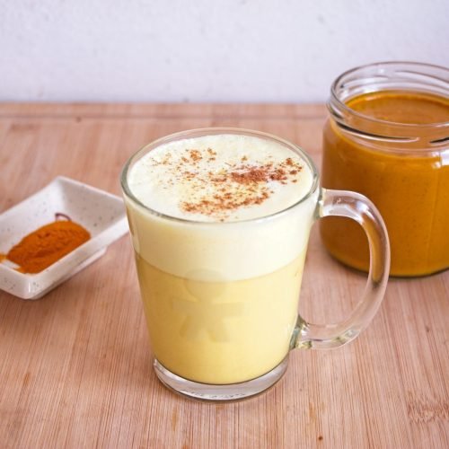 Glass mug of golden milk made with golden milk concentrate, topped with light foam and cinnamon, with a jar of concentrate in the background.