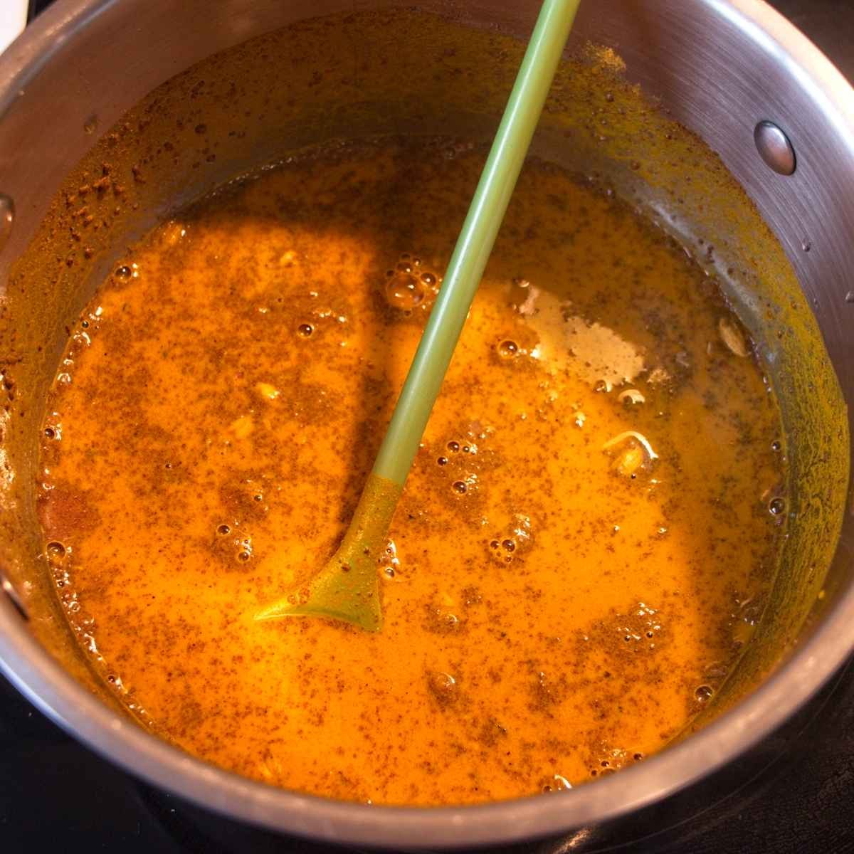 Simmering turmeric and spice mixture in a saucepan, stirred with a green spatula as it thickens and reduces.