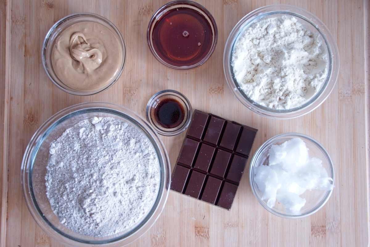 Ingredients for high protein millionaire’s shortbread arranged on a board, including oat flour, protein powder, cashew butter, maple syrup, coconut oil, and dark chocolate
