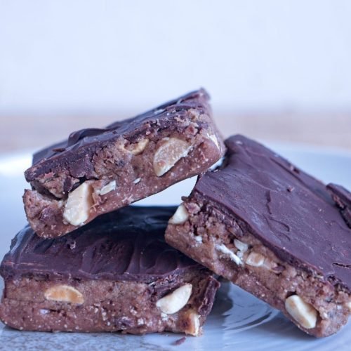 Stack of High-Protein Snickers Bites on a plate, showing the peanut nougat, date caramel, roasted peanuts, and dark chocolate top layer.