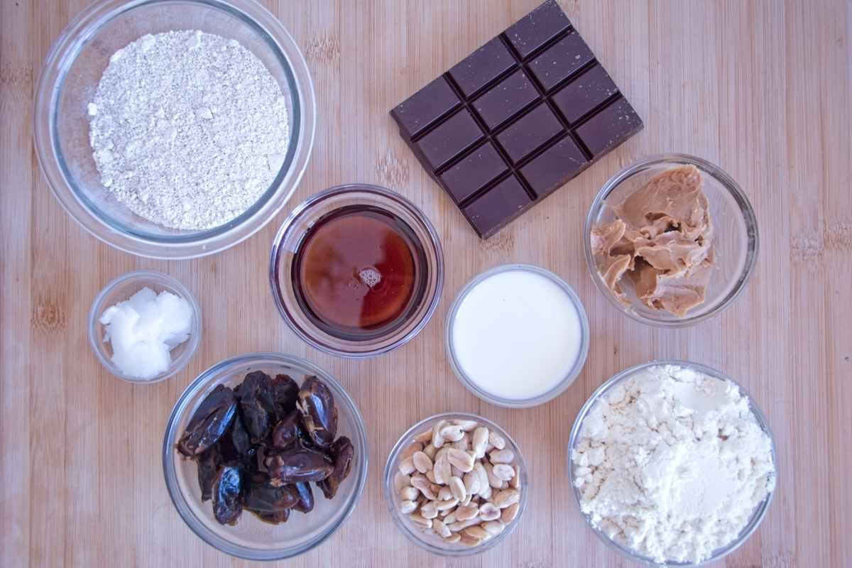 Ingredients for High-Protein Snickers Bites arranged on a board, including oat flour, whey protein, peanut butter, dates, maple syrup, milk, dark chocolate, coconut oil, and roasted peanuts.