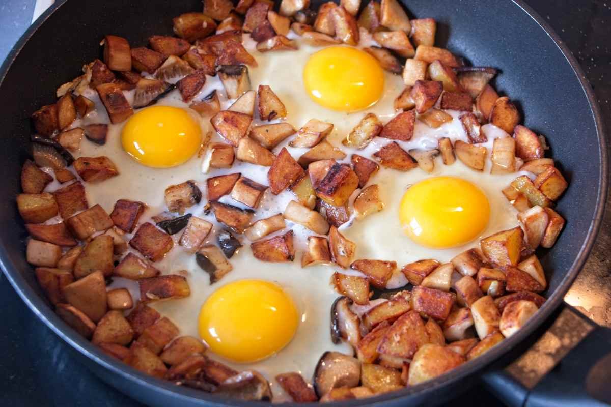 Four eggs cracked into a skillet of browned potatoes and mushrooms before cooking.