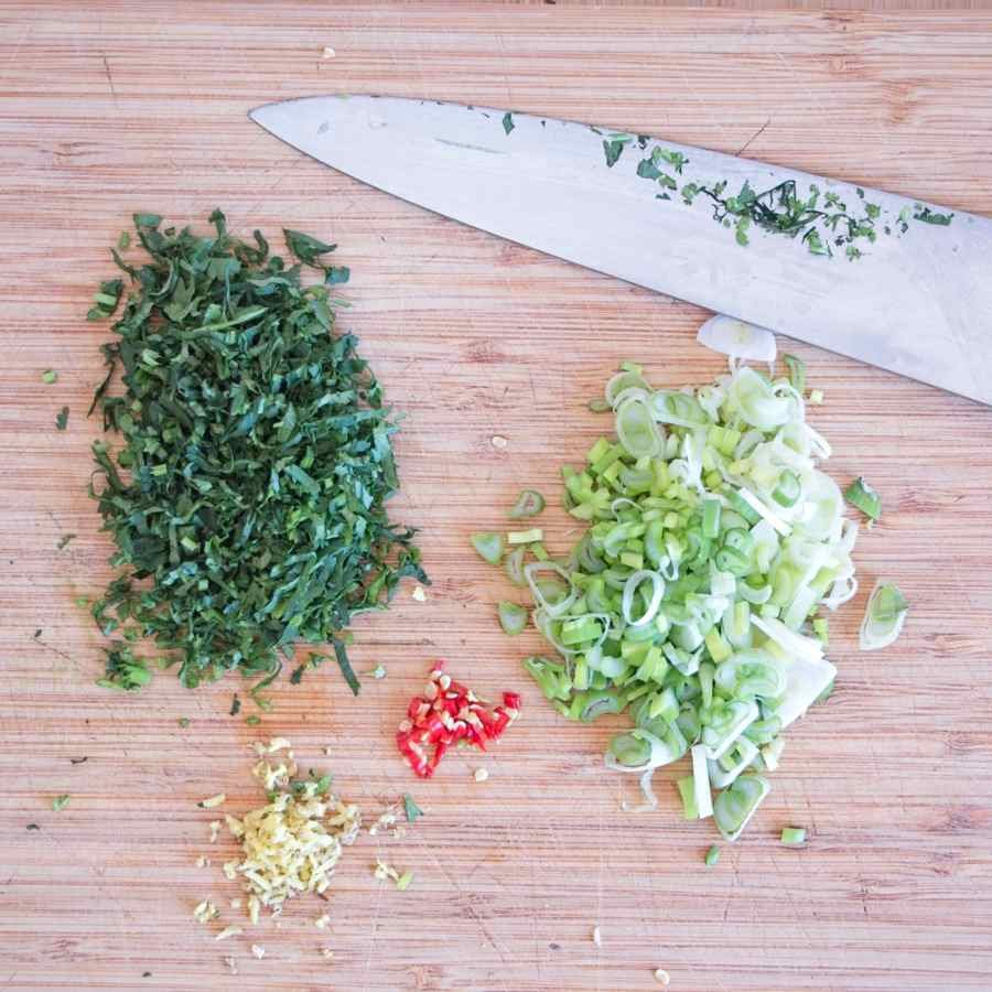 Chopped coriander, sliced spring onions, minced ginger, and red chilli prepared on a wooden board with a chef’s knife.