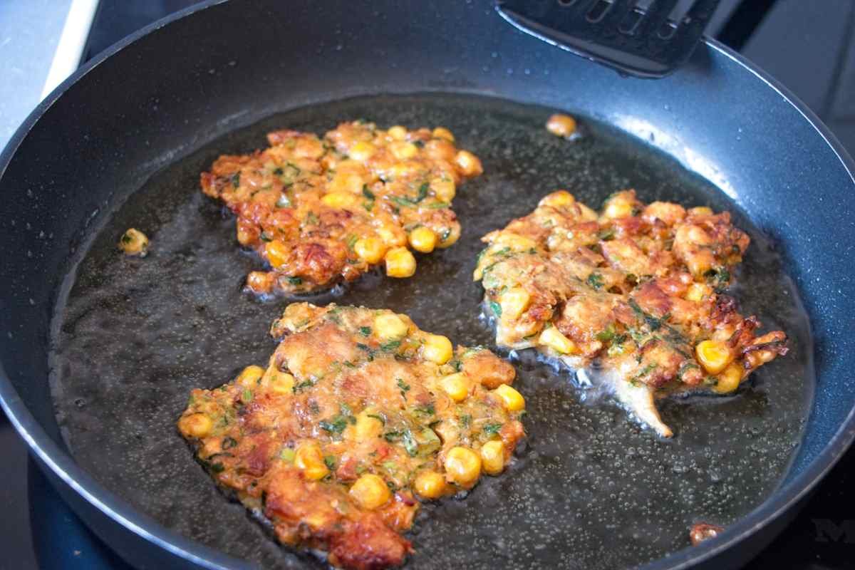 Fritters frying in oil in a pan, turning deep golden with crisp edges and visible sweetcorn pieces.