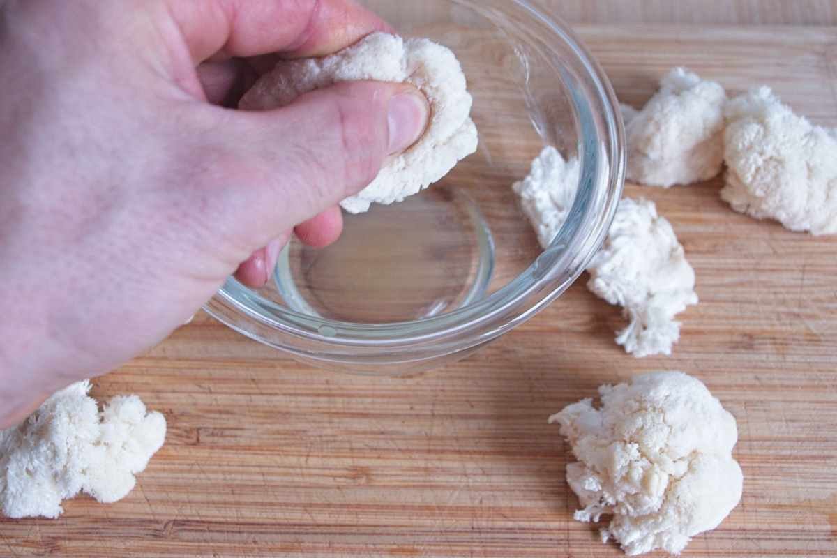 Hand squeezing excess moisture from torn lion’s mane mushroom pieces over a small glass bowl.