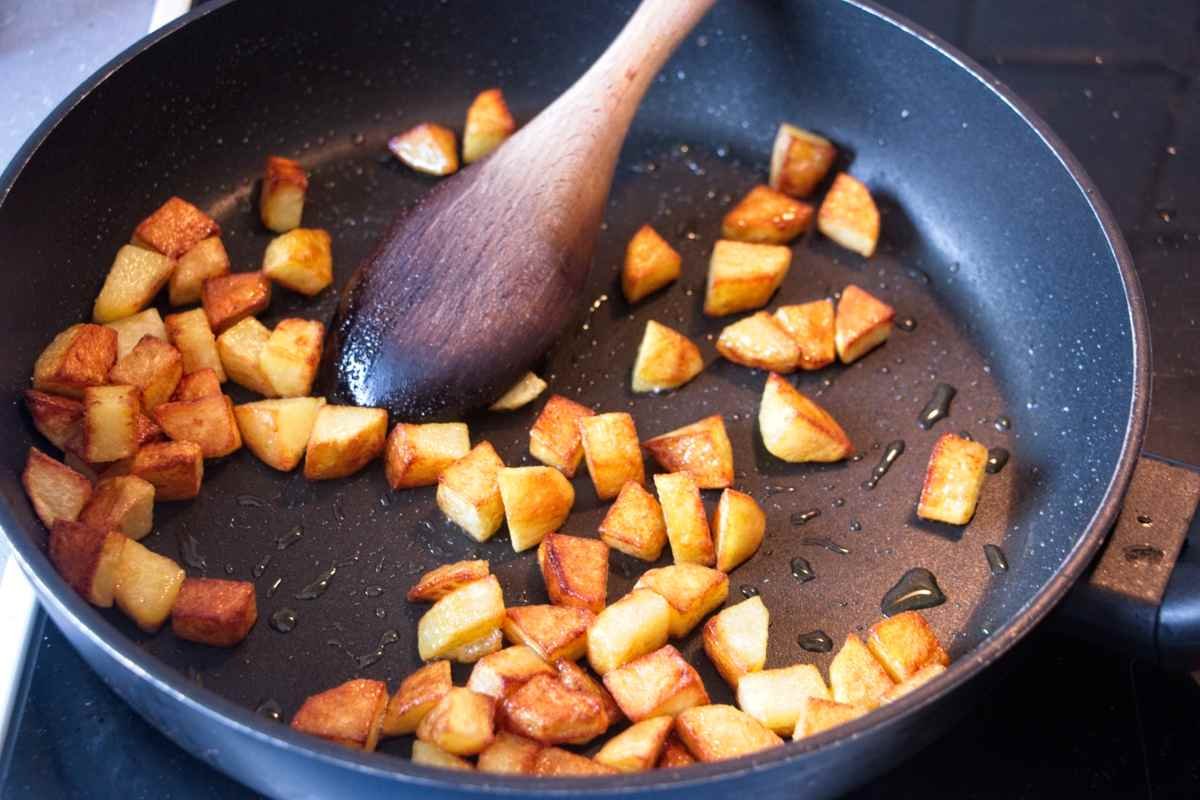 Diced potatoes browning in a skillet with oil, stirred with a wooden spoon.
