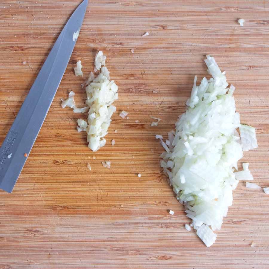 Finely chopped onion and minced garlic on a wooden board beside a chef’s knife, prepared for cooking.