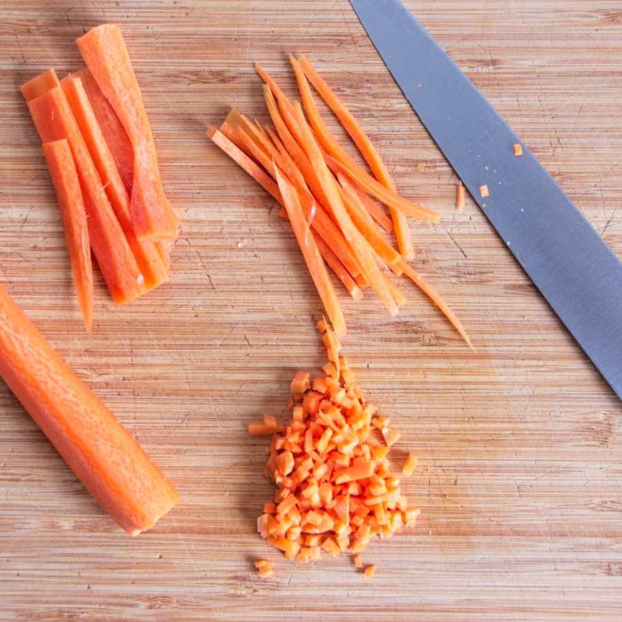Carrots sliced and finely diced on a wooden board with a chef’s knife, showing preparation for the sauce base.