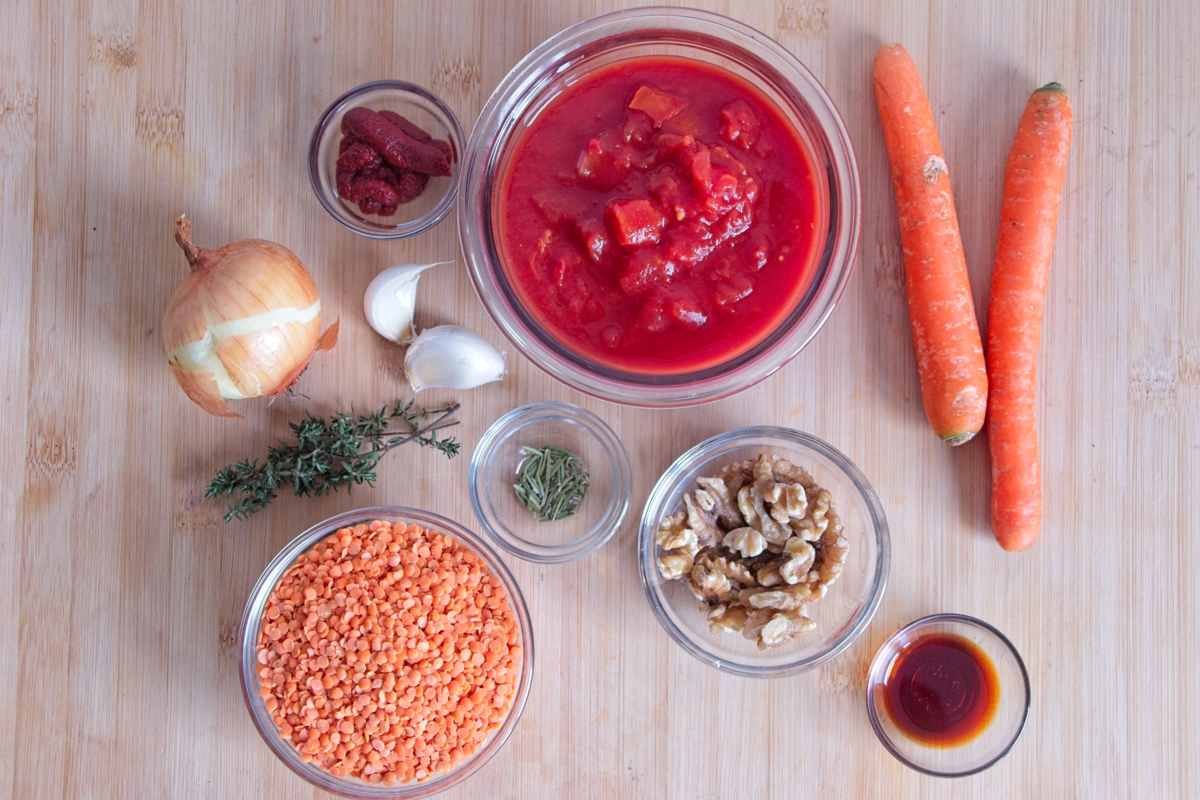 Ingredients for red lentil walnut ragu arranged on a board, including red lentils, walnuts, chopped tomatoes, onion, carrots, garlic, herbs, and tomato paste.