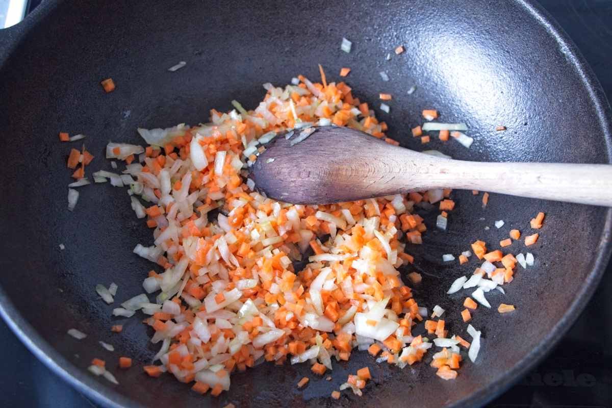 Finely diced onion and carrot sautéing in a pan with a wooden spoon to form the base of the sauce.