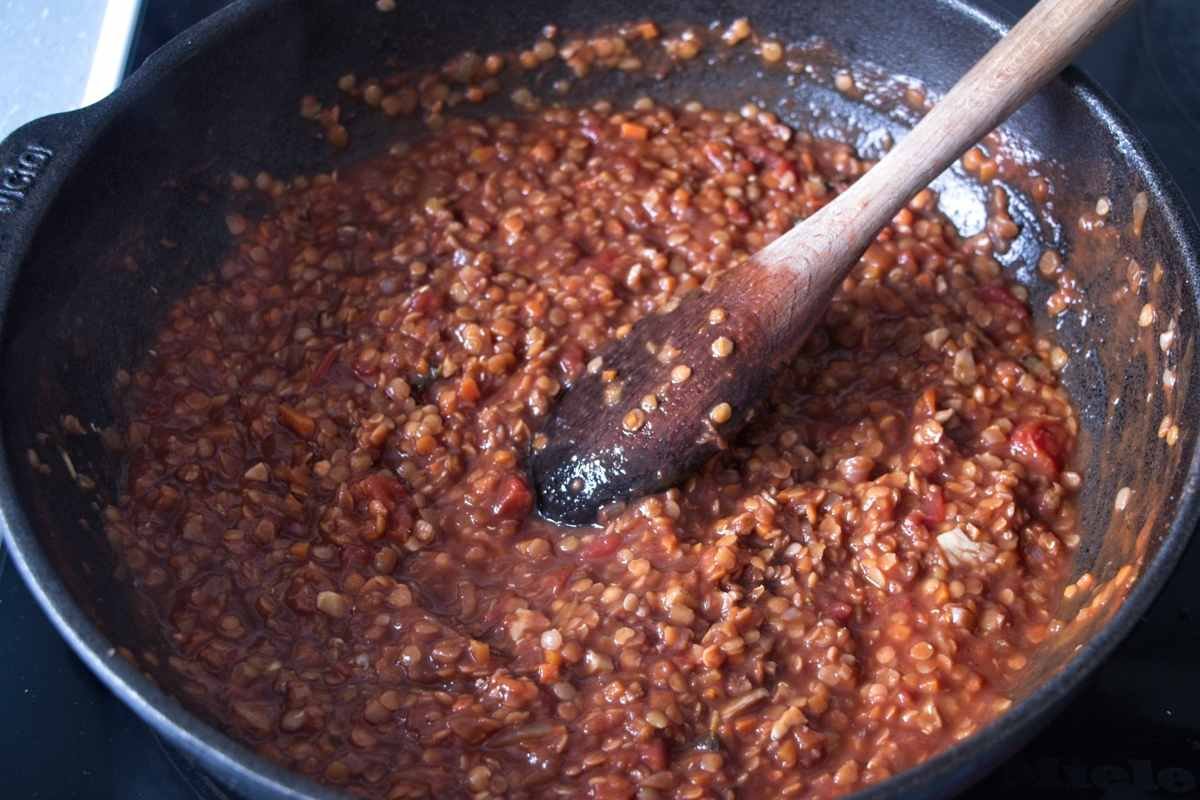 Lentil and walnut sauce simmering in a cast iron pan with a wooden spoon, showing a thick tomato base and softened lentils.