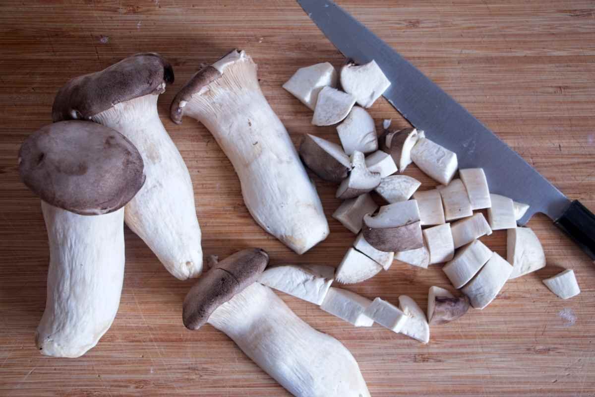 Whole and sliced king oyster mushrooms on a wooden board next to a chef’s knife.