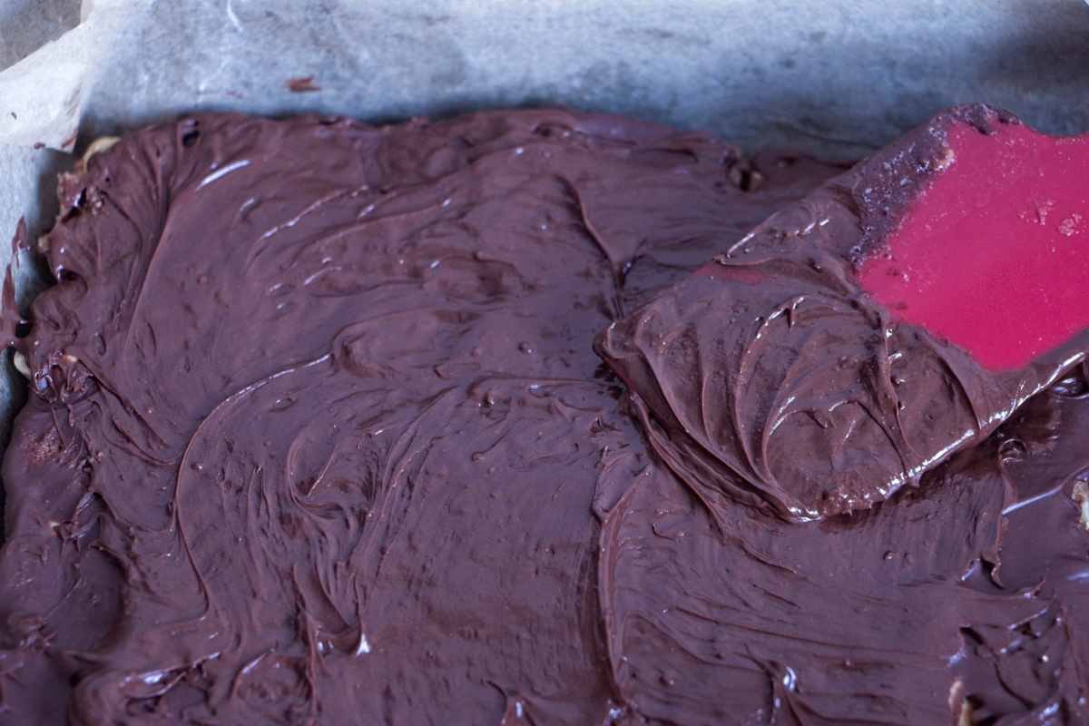 Melted dark chocolate being spread evenly over the caramel layer with a spatula in a lined tin.
