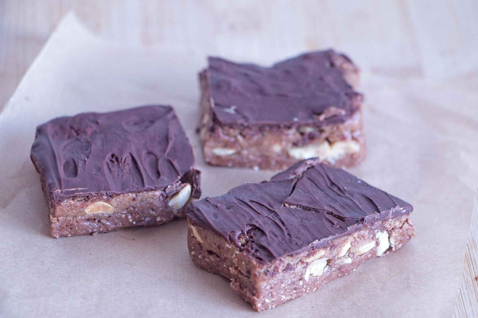 High-Protein Snickers Bites cut into squares with a peanut nougat base, date caramel layer, and dark chocolate top on parchment paper.
