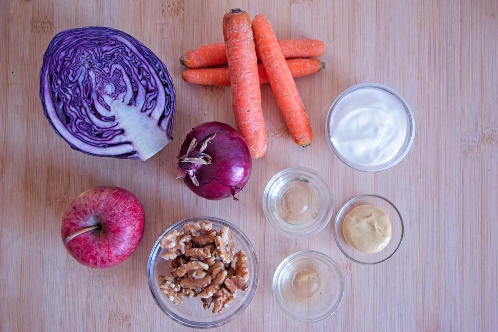 Winter coleslaw ingredients laid out on a board, including red cabbage, carrots, apple, walnuts, Greek yogurt, mustard, vinegar, and olive oil.