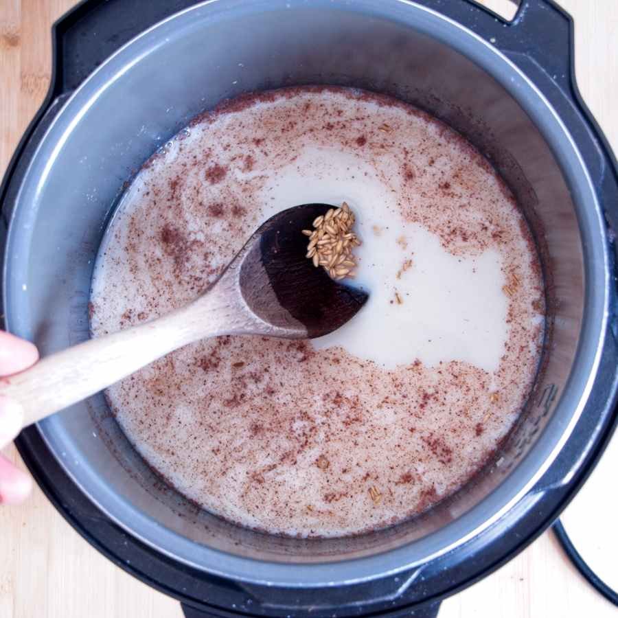 Oat groats cooking in a pressure cooker with milk, cinnamon, and a wooden spoon stirring the mixture