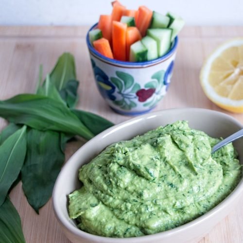 wild garlic hummus in bowl with lemon, fresh leaves, and sliced vegetables on wooden board