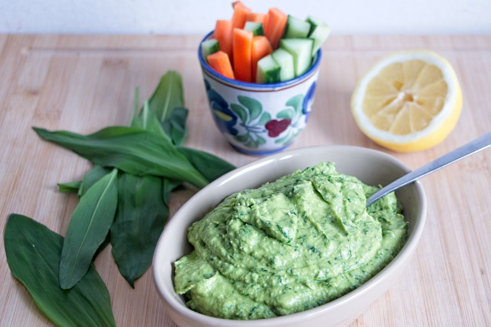 wild garlic hummus in bowl with lemon, fresh leaves, and sliced vegetables on wooden board