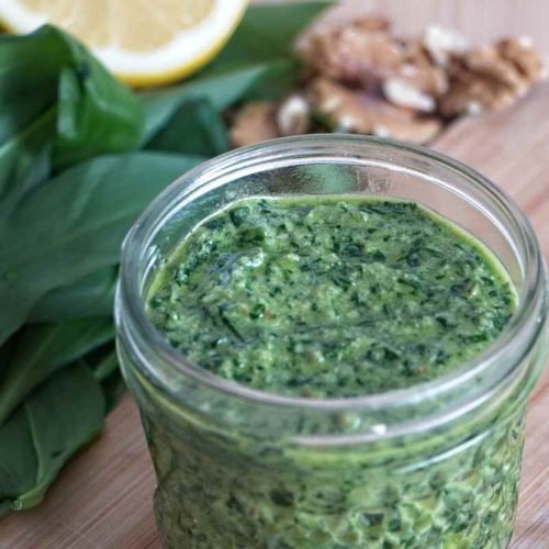 Jar of wild garlic pesto on a wooden board with fresh leaves, lemon, and walnuts in the background