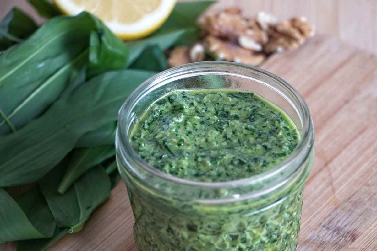Jar of wild garlic pesto on a wooden board with fresh leaves, lemon, and walnuts in the background