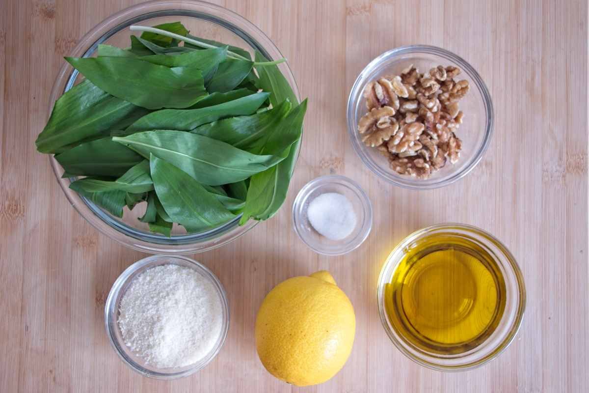 Ingredients for wild garlic pesto including fresh leaves, walnuts, parmesan, lemon, olive oil, and salt laid out on a wooden board
