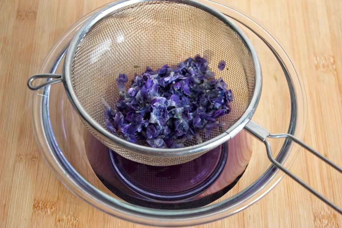 Straining violet petals from infused liquid through a sieve into a glass bowl