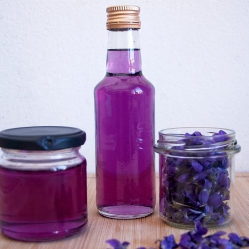 Bottle and jar of violet syrup with fresh violet petals on a wooden surface