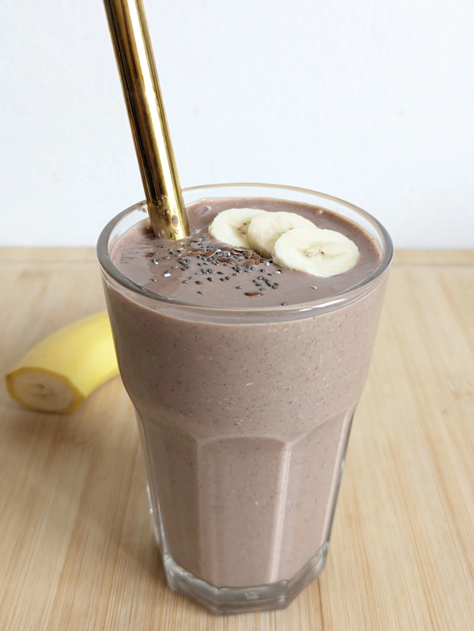 Chocolate high protein breakfast smoothie with a reusable metal straw, topped with banana slices, chia seeds and ground flaxseed in a glass.