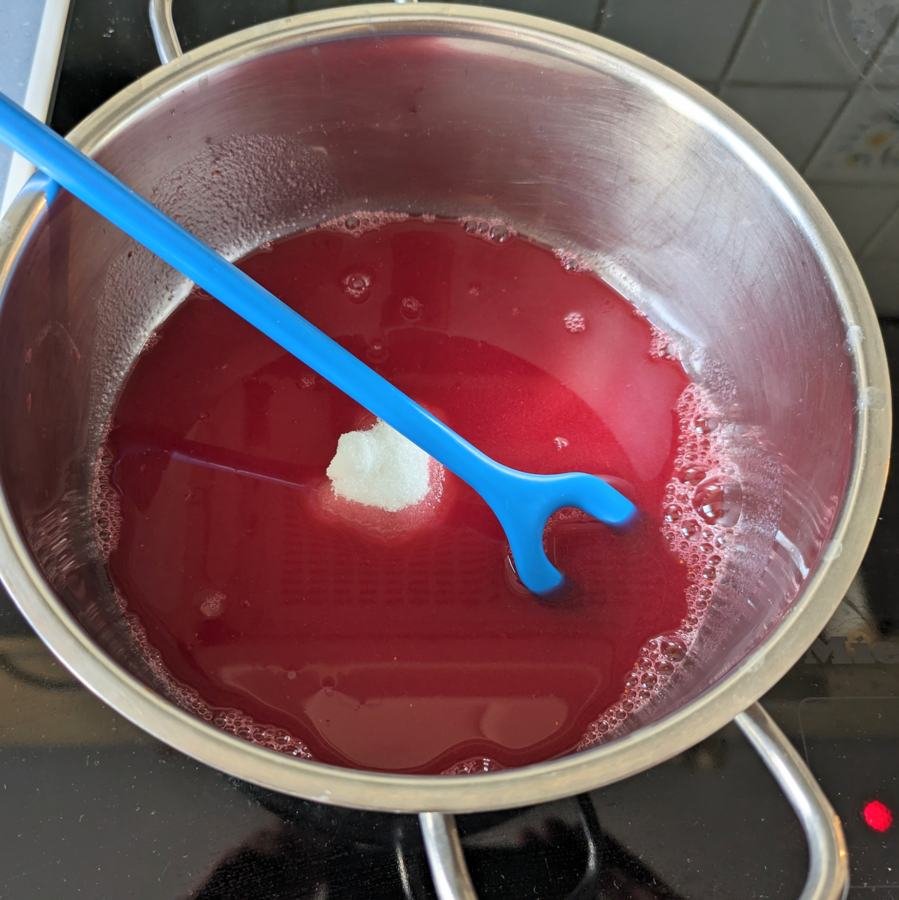 Sugar being stirred into strained lingonberry juice in a saucepan.