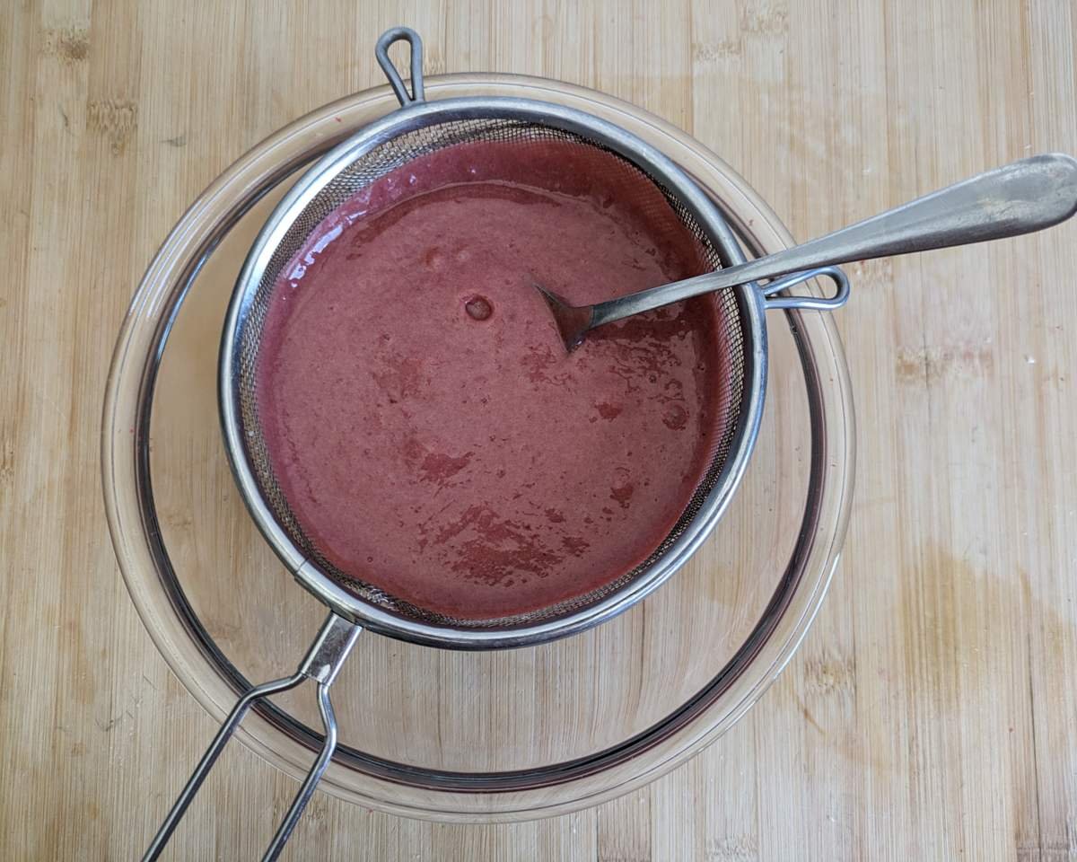 Cornelian cherry curd being pressed through a fine-mesh sieve into a bowl.