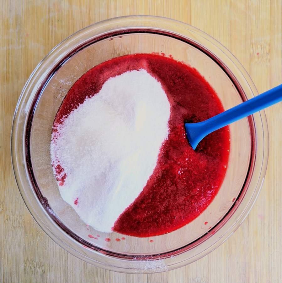 Sugar added to red fruit purée in a glass mixing bowl.