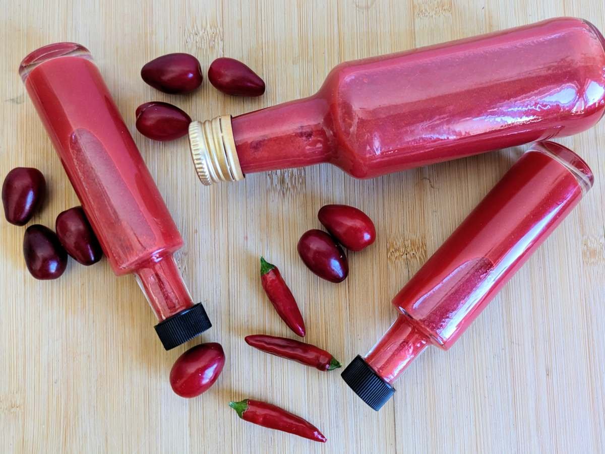 Bottles of Cornelian cherry hot sauce arranged with fresh cherries and red chillies.