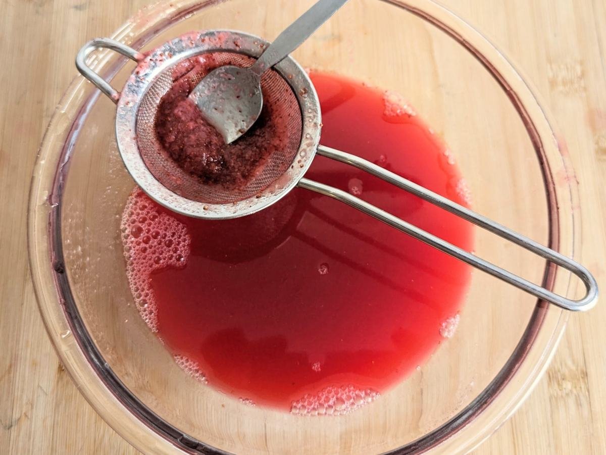 Lingonberry juice being passed through a fine-mesh sieve to remove remaining berry pulp and seeds.