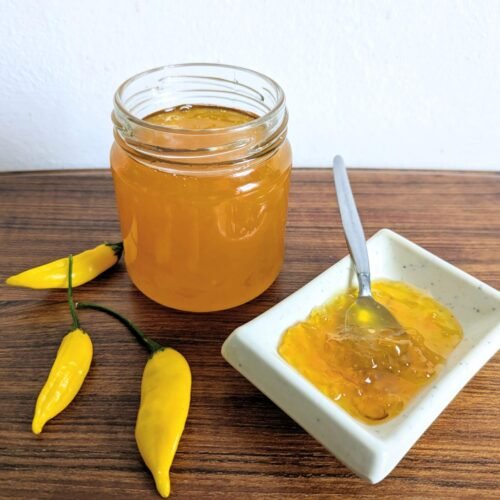 Open jar of golden Lemon Drop chilli jam beside a dish of jam and three yellow chillies.
