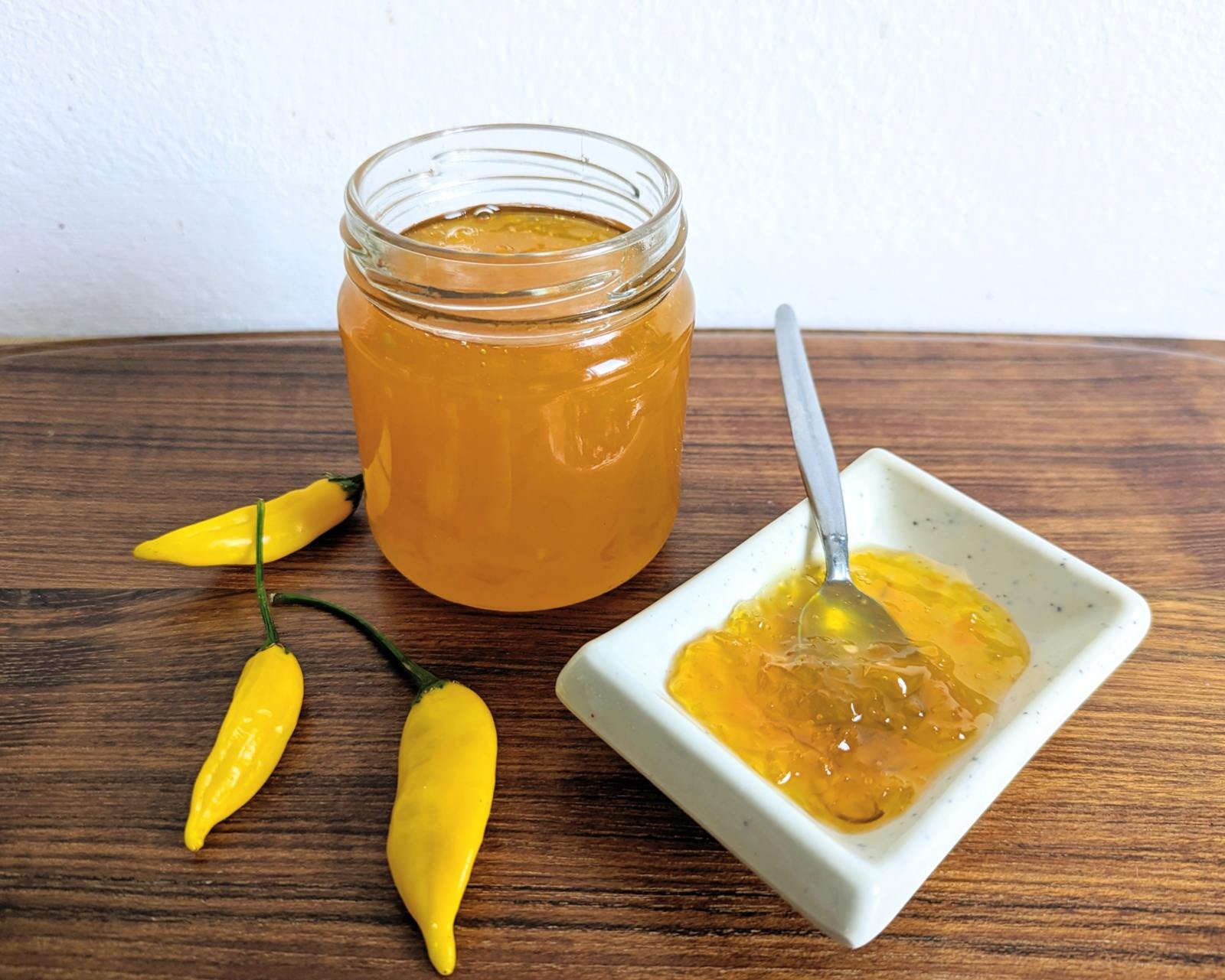 Open jar of golden Lemon Drop chilli jam beside a dish of jam and three yellow chillies.