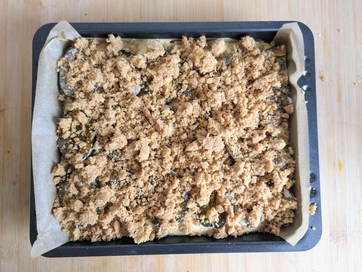 Unbaked cake topped with fresh prune plums and cinnamon streusel in a square baking tin.