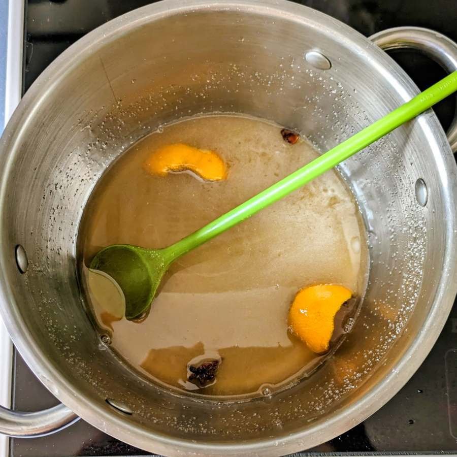 Spiced syrup with citrus peel and star anise in a stainless steel pan with a green spoon.