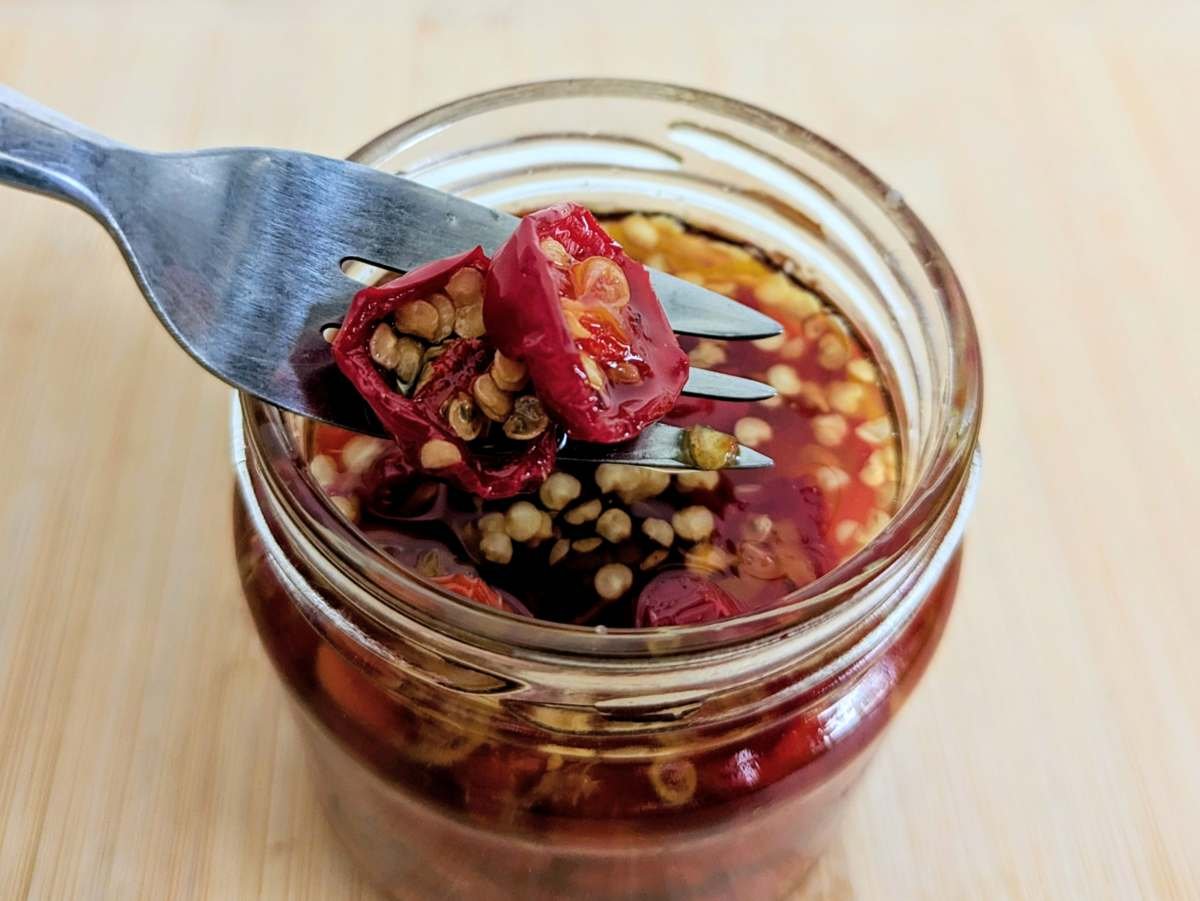 Close-up of candied red jalapenos on a fork above an open jar, with glossy chilli rings and seeds visible.