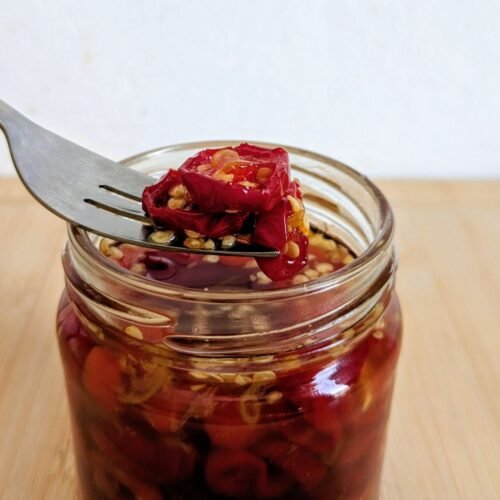 Candied red jalapenos on a fork above an open glass jar filled with chilli rings and amber syrup