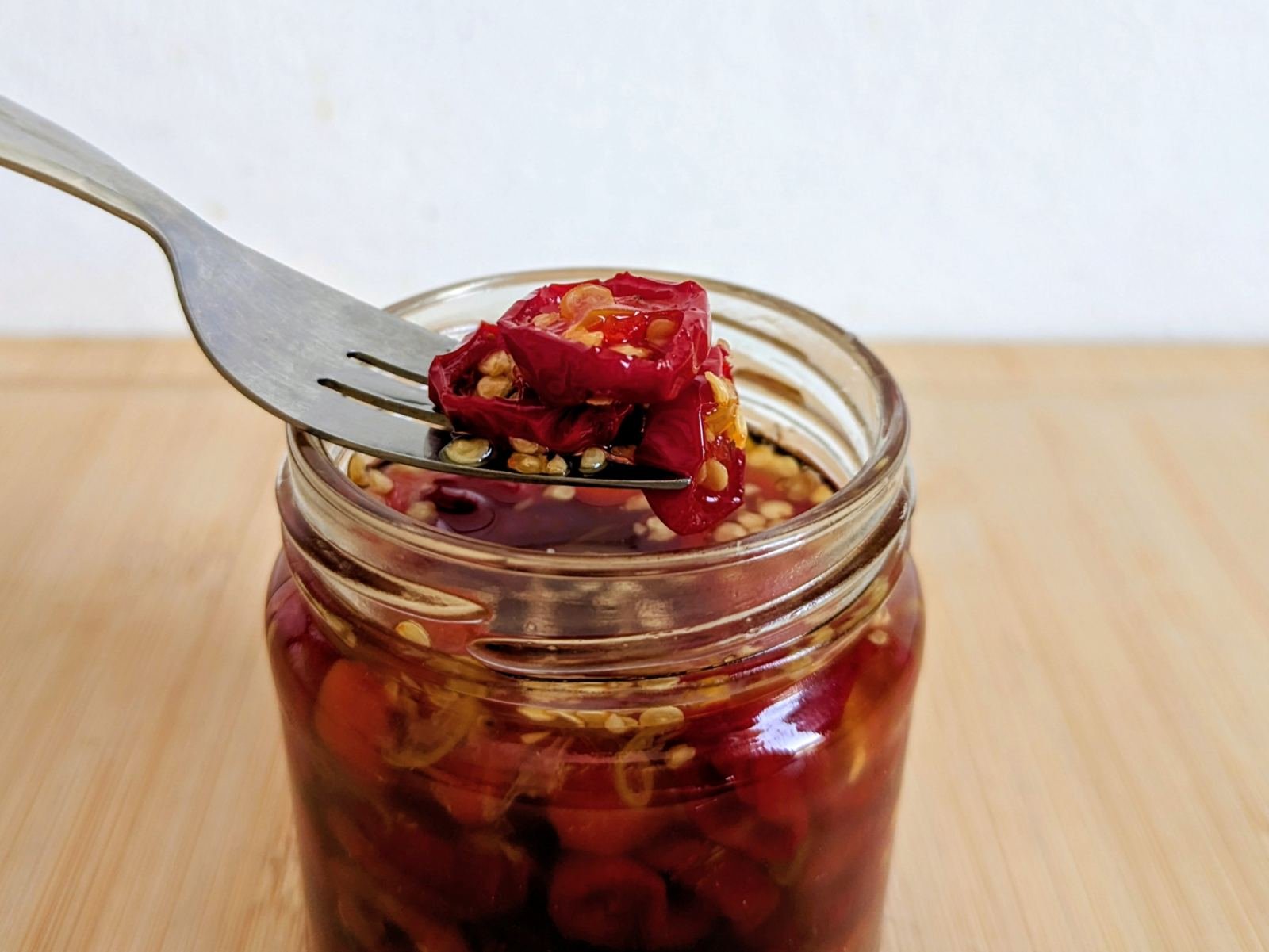 Candied red jalapenos on a fork above an open glass jar filled with chilli rings and amber syrup