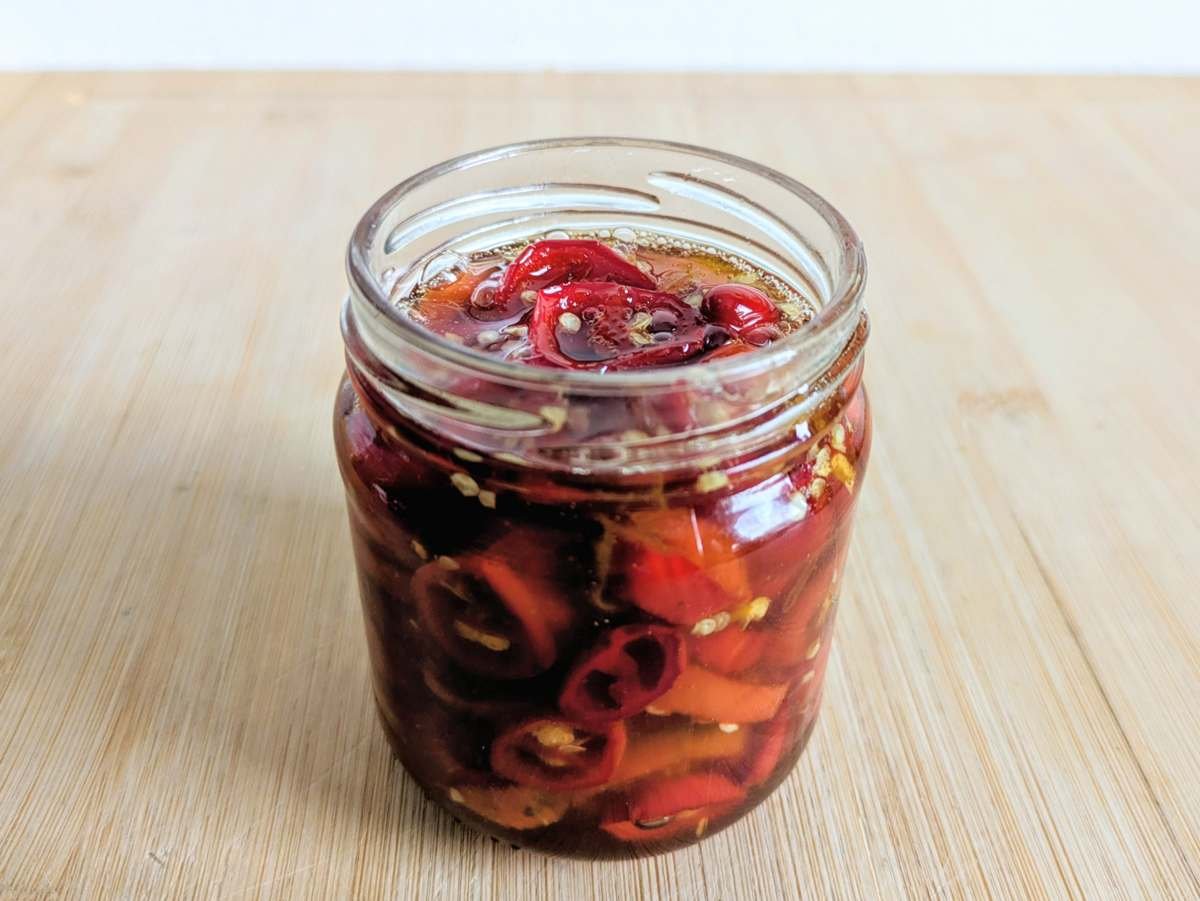 Candied red jalapenos in amber syrup in an open glass jar on a wooden board.