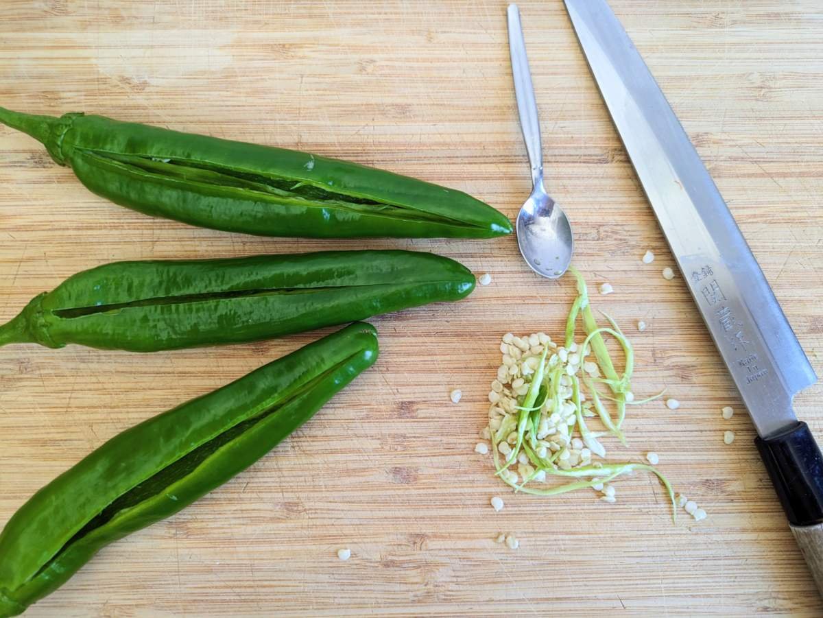 Three green peppers slit lengthways, with removed seeds and inner ribs beside a teaspoon and knife on a chopping board.