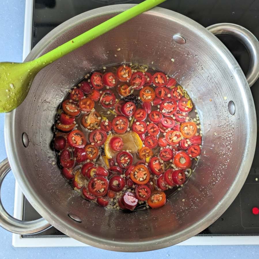 Sliced red jalapeños simmering in syrup with citrus peel and star anise in a stainless steel pan.