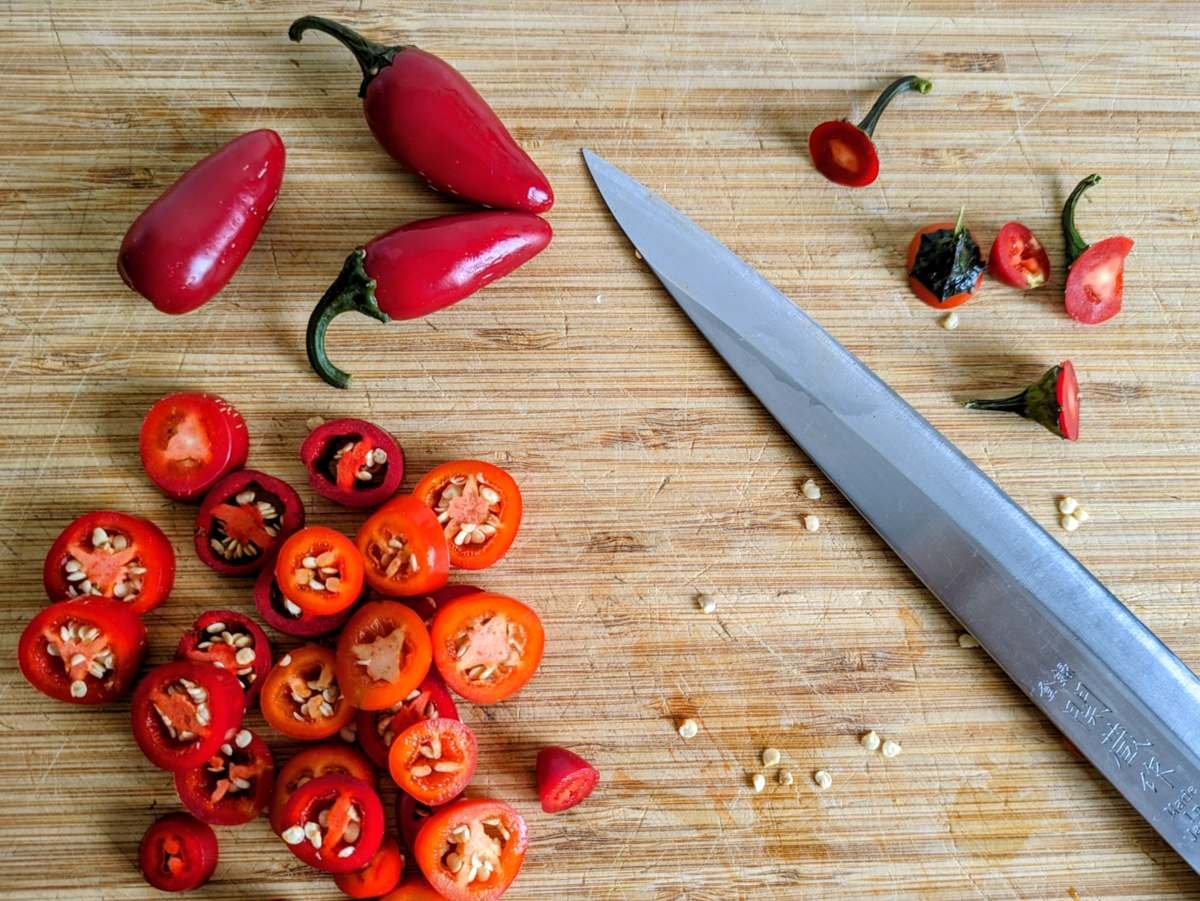 Whole red jalapeños and sliced chilli rings on a wooden chopping board beside a kitchen knife.