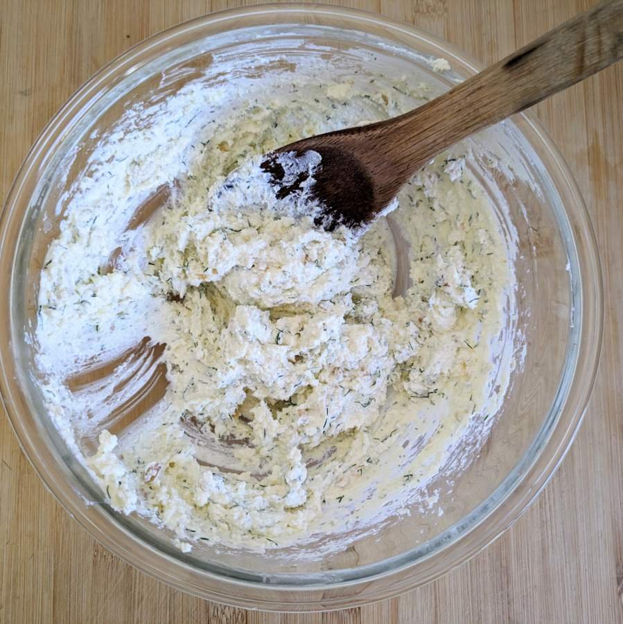 Cheese filling mixed with herbs and pine nuts in a glass bowl with a wooden spoon.
